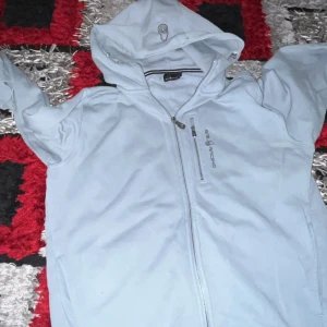 Ljusblå jacka från Sail Racing - Snygg ljusblå jacka från Sail Racing med dragkedja och huva. Perfekt för svala dagar. Jackan har en stilren design med logotyp på bröstet och praktiska fickor med dragkedja. Kom med bud 