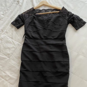 Svart klänning med spetsdetaljer - Elegant svart klänning med spetsdetaljer på ärmarna och överdelen. Klänningen har en figurnära passform och korta ärmar. Perfekt för en stilren look.