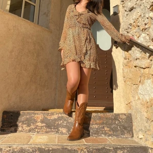 Leopardmönstrad klänning med volangdetaljer - Trendig leopardmönstrad klänning med volangdetaljer och v-ringning. Klänningen har långa ärmar och en kort längd, perfekt för en chic look. Tillverkad i ett lätt och luftigt material som ger en bekväm passform.