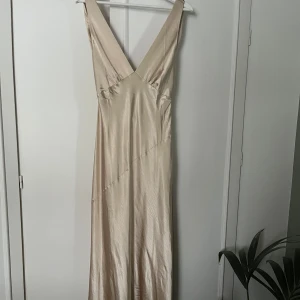 Champagnefärgad långklänning med öppen rygg - Elegant champagnefärgad långklänning i satin med djup v-ringning och öppen rygg. Klänningen har en smickrande passform och ett vackert fall, perfekt för festliga tillfällen såsom bal. 