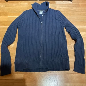 Mörkblå ribbad tröja från Hilfiger Denim - Säljer en stilren mörkblå ribbad tröja från Hilfiger Denim. Tröjan har en dragkedja framtill och en liten broderad logga på bröstet. Perfekt för kyligare dagar med sin höga krage och långa ärmar.