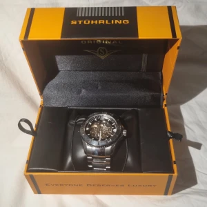 Silverfärgad klocka från Stührling - Snygg silverfärgad klocka från Stührling med synlig urtavla och robust metallarmband. Klockan kommer i en elegant orange och svart förpackning. Perfekt för den som vill ha en stilren och lyxig accessoar. Originelt köpt för 2700 men säljs för 2000. Finns ett litet fel på ringen runt glaset då den sitter lite löst. Sparsamt använd och i mycket bra skick. Cirka 5 Linus är antagna och de linksen följer med.