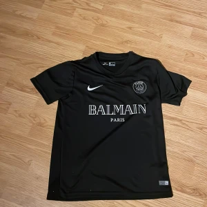 Svart fotbollströja från Nike  - Snygg svart fotbollströja från Nike med Balmain Paris-logga på bröstet. Tröjan är kortärmad och tillverkad i Dri-FIT-material för optimal komfort. Perfekt för både träning och vardag.