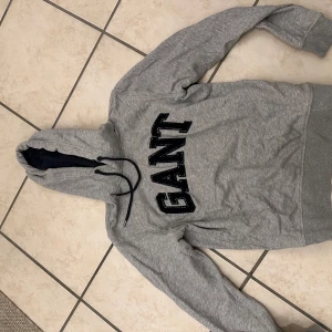 Grå hoodie från GANT - Säljer en grå hoodie från GANT med svart logotyp på bröstet. Tröjan har en klassisk design med luva 