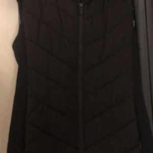 Svart quiltad väst från Zara - Säljer en stilren svart quiltad väst från Zara. Västen har wen praktisk dragkedja framtill och är perfekt för lager-på-lager under kyligare dagar.