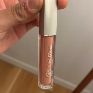 Beige läppglans med silverlock - Snyggt beige läppglans i en genomskinlig tub med ett stilrent silverlock. Perfekt för en naturlig look med lite extra glans.