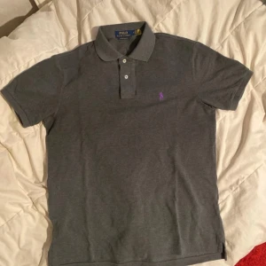 Grå pikétröja från Ralph Lauren - !knappt använda! Snygg grå pikétröja från Ralph Lauren i custom slim fit. Tröjan har en klassisk krage med knappar och en liten broderad logga i lila på bröstet. Perfekt för en stilren look.