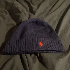 Mörkblå stickad mössa från Ralph Lauren - Snygg mörkblå stickad mössa från Ralph Lauren med det ikoniska röda broderade logotypen framtill. Perfekt för kyliga dagar och ger en stilren look.