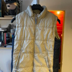 Beige dunväst från Cross Way - Säljer en beige dunväst från Cross Way med dragkedja och hög krage. Västen har två praktiska fickor med dragkedja framtill och ett broderat märke på baksidan av kragen. Perfekt för kyliga dagar. Barnstorlek 146 men passar både en small och xs
