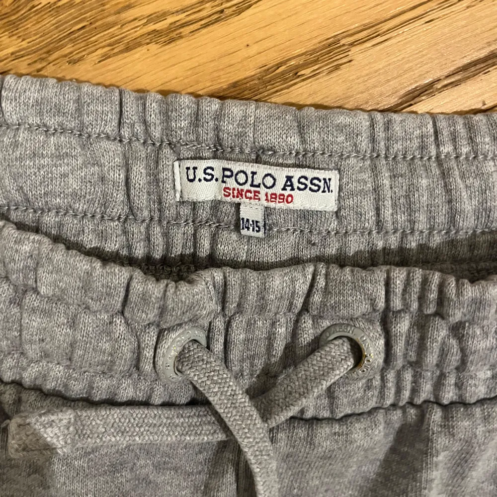 Snygga grå mjukisbyxor från U.S. Polo Assn. med elastisk midja och snörning. Byxorna har en cool randig detalj i rött, vitt och svart längs sidorna samt en ficka. Perfekta för en avslappnad stil.. Farkut & Housut.