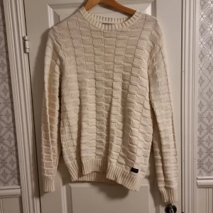 Vit stickad tröja från GANT - Säljer en stilren vit stickad tröja från GANT med ett diskret rutmönster. Tröjan har långa ärmar och ribbade muddar vid ärmslut och nederkant. Priside' 250kr