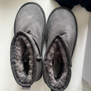 Grå UGG boots med pälsfoder - Säljer ett par grå UGG boots med mjukt pälsfoder. Skorna har en klassisk design med rund tå och en liten dragflik baktill. Perfekta för kalla dagar. 