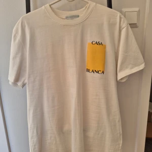 Vit t-shirt från Casablanca - Vit t-shirt från Casablanca med ett stort gult tryck och texten 'CASA BLANCA' på både fram- och baksidan. T-shirten har en klassisk rund hals och korta ärmar. Perfekt för en stilren och modern look. Helt ny