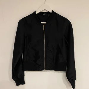 Svart bomberjacka från Heartquake - Tunn vindkacka. Snygg svart bomberjacka från Heartquake med dragkedja framtill. Jackan har en klassisk design med långa ärmar och ribbade muddar vid ärmslut och nederkant. Perfekt för en stilren look.