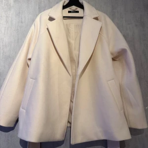 Beige kappa från Gina Tricot - Snygg beige kappa från Gina Tricot med klassisk design. Jackan har en stilren look med knytbälte i midjan och två fickor framtill. Perfekt för att hålla stilen under kyligare dagar.