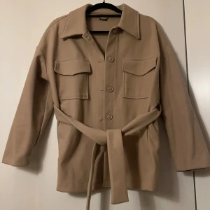 Beige jacka  med knytbälte - Snygg beige overshirt med knytbälte och stora knappar. Jackan har långa ärmar och två stora bröstfickor med lock. Passar även en s/m så den är stor i storleken