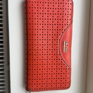 Orange plånbok från Hugo Boss - Snygg röd plånbok från Boss med perforerat mönster och dragkedja. Perfekt för att hålla ordning på kort och kontanter. Stilren design med gott om fack inuti.