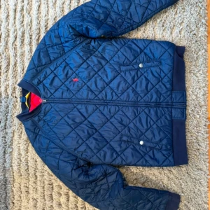 Blå quiltad jacka från Polo Ralph Lauren - Snygg blå quiltad jacka från Polo Ralph Lauren med dragkedja och tryckknappar. Jackan har en klassisk design med ribbade muddar och en liten logotyp på bröstet. Vår/höst jacka inköpt 2020, ordinarie pris 2499 kr. Stl L, 14-16 Y. 