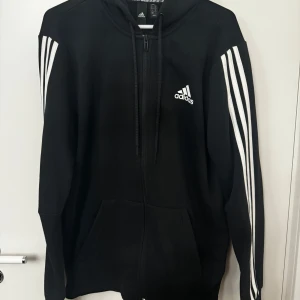 Svart hoodie från Adidas - Säljer en svart hoodie från Adidas med klassiska vita ränder längs ärmarna och en dragkedja framtill. Perfekt för en sportig look eller avslappnade dagar. Den har en justerbar huva och två fickor framtill. Storlek M. I bra skick 