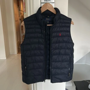Polo Ralph Lauren väst - Snygg marinblå dunväst från Polo Ralph Lauren med dragkedja och broderad logga på bröstet. Perfekt för lager-på-lager under kyliga dagar. Västen är i toppskick och nypris på västen ligger runt 3500 kr. Västen passar dig mellan 155-160 cm lång. Hör av dig vid minsta fråga.