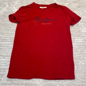 Röd t-shirt från Pepe Jeans - Snygg röd t-shirt från Pepe Jeans med blå logotyp på bröstet. Perfekt för en casual look. Tillverkad i mjukt material för extra komfort.