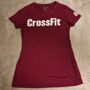 Vinröd CrossFit t-shirt från Reebok - Snygg vinröd t-shirt från Reebok med CrossFit-tryck i vitt på framsidan. Tillverkad i ett bekvämt material med Speedwick-teknologi för att hålla dig torr under träning. Perfekt för dig som älskar att träna och vill ha en stilren look.