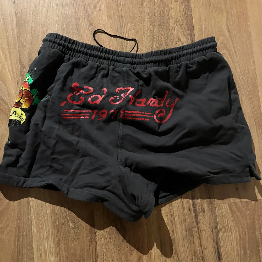 Säljer ett par svarta shorts från Ed Hardy x Urban Outfitters med färgglatt tryck och text. De har en elastisk midja med dragsko för justerbar passform. Perfekta för en avslappnad stil.. Shortsit.