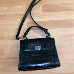 Svart krokodilpräglad handväska - Snygg svart handväska med krokodilpräglat mönster och en praktisk axelrem. Väskan har ett elegant spänne framtill och är perfekt för att bära dina nödvändigheter med stil.