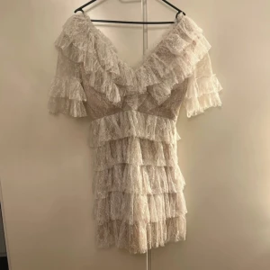 Vit spetsklänning från By Malina - Elegant vit spetsklänning från By Malina med volanger och v-ringad design. Klänningen har korta ärmar och flera lager av spets som ger en romantisk känsla. Min student klänning från 2024. Använd en gång.
