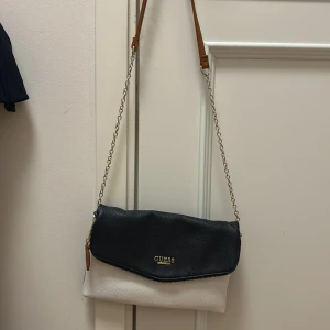 Mörk blå och vit axelväska från Guess - Snygg axelväska från Guess i mörkblått och vitt med gulddetaljer. Väskan har en kedjerem och en praktisk dragkedja för säker förvaring. Perfekt för att bära med sig det viktigaste med stil.