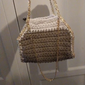 Grå/vit virkad väska med guldkedja - Snygg virkad väska i grå och vitt med en elegant guldkedja. Perfekt för att ge din outfit en unik touch. Väskan har en rymlig design och är handgjord med ett vackert mönster.