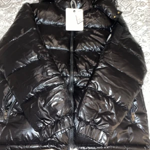 Svart pufferjacka från Moncler - Säljer en snygg svart pufferjacka från Moncler med glansig finish. Jackan har en dragkedja framtill och praktiska fickor med dragkedjor. Perfekt för kyliga dagar med sin quiltade design.