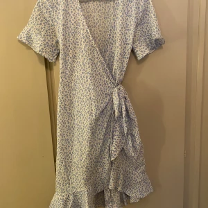 Blåmönstrad omlottklänning - Charmig omlottklänning med blått leopardmönster på vit bakgrund. Klänningen har korta ärmar och en volangdetalj vid fållen. Perfekt för en somrig look med sin lätta och luftiga design.