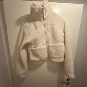 Vit teddyjacka  med dragkedja - Mysig vit teddyjacka med hög krage och dragkedja framtill. Jackan har långa ärmar och två stora fickor. Säljer denna trendiga teddyjacka från H&M i storlek s. Använd 3 gånger och i mycket bra skick!