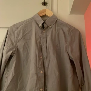 Grå skjorta från Ralph Lauren - Säljer en klassisk grå skjorta från Ralph Lauren i storlek L. Skjortan har långa ärmar och knappar framtill samt en button-down krage. Perfekt för dig som är är 14 till 16 år