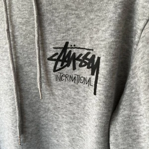 Grå hoodie från Stüssy - Säljer en grå hoodie från Stüssy med deras ikoniska logga både fram och bak. Tröjjan är i gott skick!