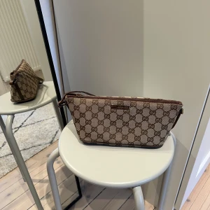Gucci pochette bag - Brun Gucci väska med klassiskt GG-mönster och dragkedja. Väskan har en kort axelrem och en liten metallbricka med Gucci-logotypen på framsidan. Den är väl använd som bilderna kan visa men mycket kvar att ge! Lilla ”flärpen” på dragkedjan har lossnat men den finns kvar om man vill limma dit den :) Tyget är inte skadat. Köpt på The Vintage Bar, kvitto finns