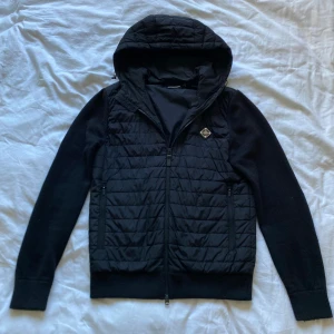 Svart hybridjacka från J.Lindeberg - Snygg svart hybridjacka från J.Lindeberg med quiltad design och huva. Slutsåld nästan överallt och är väldigt svårt att hitta. Mycket bra skick och är perfekt till våren. Original priset på jackan var 3200kr. 