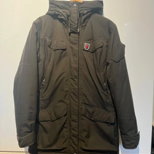 Fjällräven Jacka (Nuuk Parka) - Säljer en grön parkajacka från Fjällräven med huva och dragkedja. Jackan har flera praktiska fickor och en diskret logga på bröstet. Perfekt för kyligare väder! Jackan är i bra skick o använd ett fåtal gånger. Pälsen är avtagbar och nypris ca 6500kr.