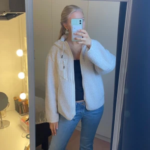 Vit teddyjacka - Mysig vit teddyjacka med dragkedja och hög krage. Jackan har en bröstficka med dragkedja och långa ärmar. Perfekt för kyligare dagar när du vill hålla dig varm och bekväm. Passar mig som vanligtvis bär storlek S