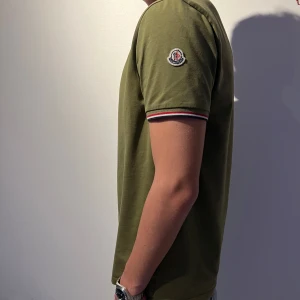 Moncler t shirt  - Snygg olivgrön t-shirt från Moncler med korta ärmar och en klassisk passform. T-shirten har Monclers logga på ärmen och randiga detaljer vid ärmsluten. Perfekt för en stilren och avslappnad look.