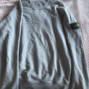 Grå sweatshirt från Stone Island - Säljer en stilren grå sweatshirt från Stone Island med deras ikoniska märke på ärmen. Tröjan har en rund halsringning och är perfekt för en avslappnad look. Passar bra till både jeans och joggers.
