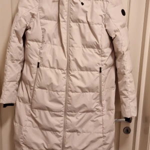 Beige dunjacka från áhkká - Säljer en stilren beige dunjacka från áhkká. Jackan har en lång design med dragkedja framtill och praktiska fickor. Perfekt för kyliga dagar med sin varma och bekväma passform.
