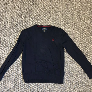 Mörkblå tröja från Polo Ralph Lauren - Säljer en stilren mörkblå tröja från Polo Ralph Lauren med röd logga på bröstet. Tröjan är tillverkad i bomull och har långa ärmar samt ribbade muddar vid ärmslut och nederkant. Perfekt för en klassisk look. Storlek M i barnstorlek och passar ungefär 12 år, runt 155 skulle jag säga 