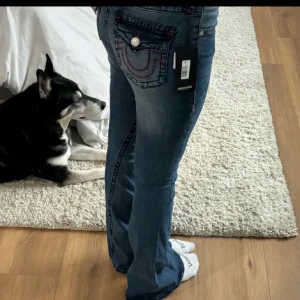 Blå jeans från True Religion - Snygga blå jeans från True Religion med bootcut-stil. De har röda sömmar och knappar på bakfickorna. Modellen heter Joey och har en midwaist-passform. Perfekta för en avslappnad look. Skriv om frågor eller bilder. 🥰