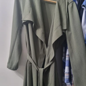 Olivgrön trenchcoat - Snygg olivgrön trenchcoat med knytband i midjan. Jackan har långa ärmar och en elegant krage som ger en stilren look. Perfekt för höst och vår.