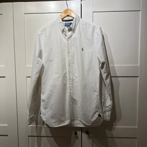 Vit skjorta från Polo Ralph Lauren - Säljer en klassisk vit skjorta från Polo Ralph Lauren i slim fit. Skjortan har långa ärmar och är dekorerad med det ikoniska broderade polospelar-emblemet på bröstet. Perfekt för en stilren look.