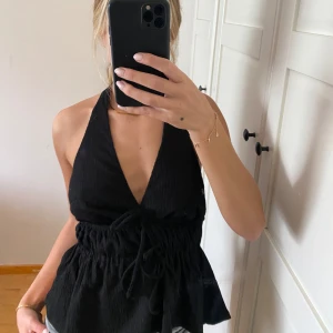 Svart halterneck topp - Snygg svart halterneck topp med knytning i midjan och volangdetaljer. Perfekt för en stilren look. Tillverkad i ett mjukt material som ger en bekväm passform.