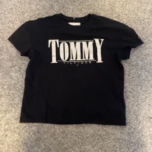 Svart t-shirt från Tommy Hilfiger - Snygg svart t-shirt från Tommy Hilfiger med stort tryck på framsidan. Perfekt för en casual look. Tillverkad i mjukt material för extra komfort.