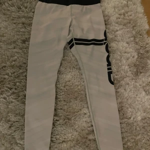 Grå leggings med svart mönster - Snygga grå leggings med ett svart grafiskt mönster längs benet. De har en elastisk midja för bekväm passform och är perfekta för en avslappnad stil. Materialet verkar vara mjukt och stretchigt.